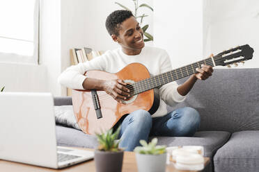 Frau spielt Gitarre auf Couch mit Laptop auf Tisch - JCZF01124