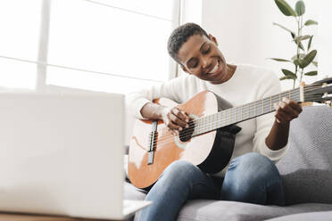 Frau spielt Gitarre auf Couch mit Laptop auf Tisch - JCZF01125