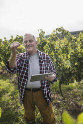Glücklicher älterer Landwirt mit Tablet-PC und Weintrauben im Weinberg - UUF27370