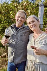 Smiling mature couple holding wineglasses in front of vineyard on sunny day - ZEDF04897