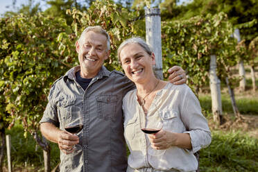 Smiling mature man and woman holding wineglasses in front of vineyard - ZEDF04899