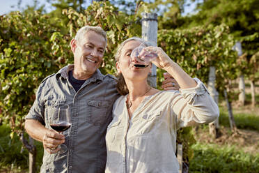 Smiling mature man holding wineglass looking at woman drinking wine in front of vineyard - ZEDF04900