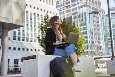 Freelancer with laptop sitting on seat in front of buildings - VEGF06004