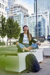 Woman with eyes closed meditating on seat in front of buildings - VEGF06005