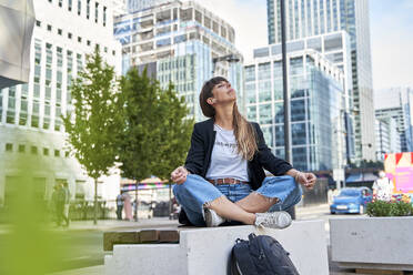 Woman with eyes closed meditating on seat in front of buildings - VEGF06006