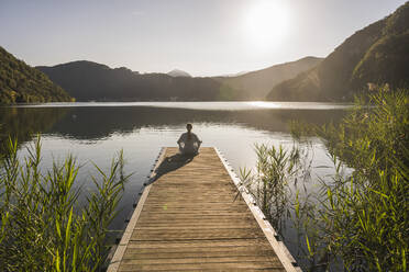 Ältere Frau meditiert auf einem Steg über einem See bei einer Bergkette - UUF27424