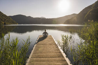 Mature woman meditating on jetty over lake by mountain range - UUF27424