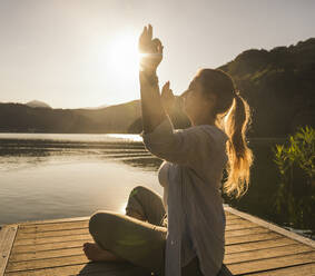 Mature woman meditating with arms raised on jetty by lake - UUF27428