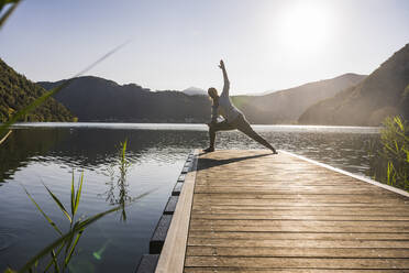 Frau übt mit erhobener Hand auf einem Steg über einem See in den Bergen - UUF27429