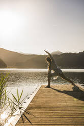 Mature woman practicing yoga on jetty over lake at vacation - UUF27431