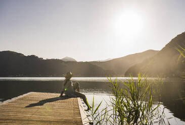 Frau mit Hut sitzt auf einem Steg über einem See in den Bergen - UUF27440