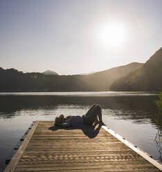 Woman lying on jetty over lake by mountains - UUF27442