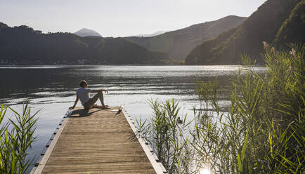 Mature man with laptop looking at lake and mountains from jetty - UUF27453