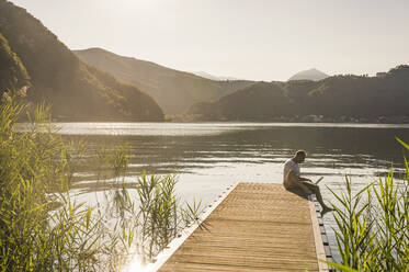 Mature man using laptop on jetty over lake by mountains - UUF27454