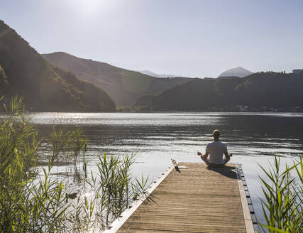 Mann mit Laptop meditiert auf Steg über See - UUF27455
