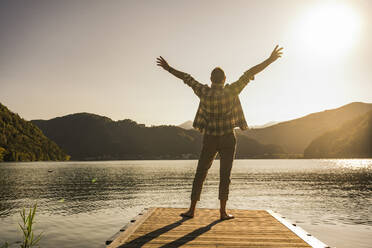 Mature man standing with arms raised on jetty by mountains - UUF27460