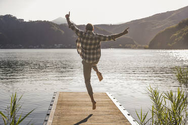 Mature man jumping on jetty over lake at vacation - UUF27462