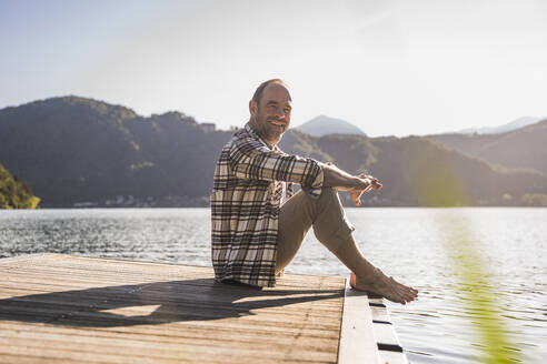 Lächelnder Mann, der im Urlaub auf einem Steg über einem See sitzt - UUF27464