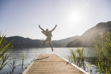 Happy mature man jumping on jetty over lake - UUF27470