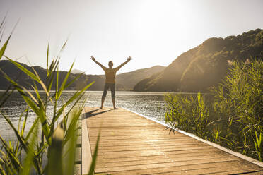 Mature man standing with arms raised on jetty at vacation - UUF27473