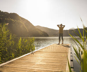 Mature man with hands behind head on jetty over lake - UUF27474