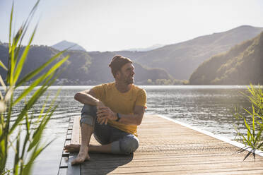 Smiling man with knit hat sitting on jetty over lake - UUF27475