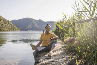 Älterer Freiberufler mit Laptop auf dem Steg am See - UUF27482