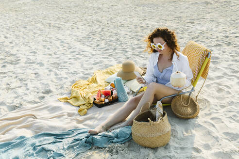 Lächelnde Frau mit Blumen-Sonnenbrille und Smartphone am Strand - TYF00446