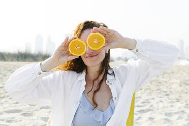 Smiling woman covering eyes with oranges at beach on sunny day - TYF00447