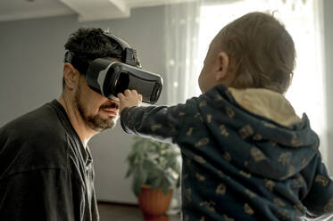 Baby boy looking at father wearing virtual reality simulator at home - ANAF00079