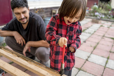 Smiling man looking at happy son painting wooden bench - ANAF00085