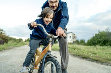 Man assisting son to ride bike on road - ANAF00091