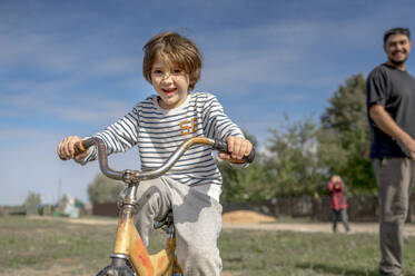 Lächelnder Junge fährt mit seinem Vater an einem sonnigen Tag Fahrrad - ANAF00098