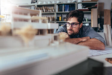 Nachdenklicher männlicher Architekt im Büro, der ein Hausmodell betrachtet. Junger Mann arbeitet an einem neuen Bauprojekt. - JLPSF02444