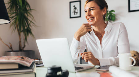 Woman sitting at her desk in office holding a pen. Smiling business woman with her laptop computer on her office desk. - JLPSF02455