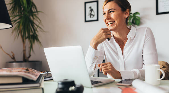 Frau sitzt an ihrem Schreibtisch im Büro und hält einen Stift. Lächelnde Geschäftsfrau mit ihrem Laptop-Computer auf ihrem Bürotisch. - JLPSF02455
