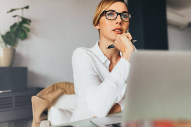 Nachdenklich blickende Geschäftsfrau, die an ihrem Schreibtisch im Büro sitzt und einen Stift in der Hand hält; Frau, die mit ihrem Laptop auf dem Tisch wegschaut und mit der Hand ihr Kinn berührt. - JLPSF02456