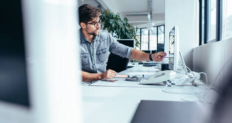 Young businessman putting post-it note on computer monitor. Caucasian male working at his desk in modern office. - JLPSF02467