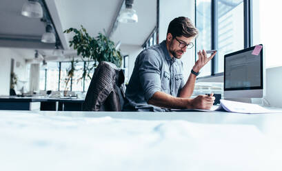 Businessman holding smart phone and working in office. Young caucasian man talking on speaker phone at his desk. - JLPSF02468