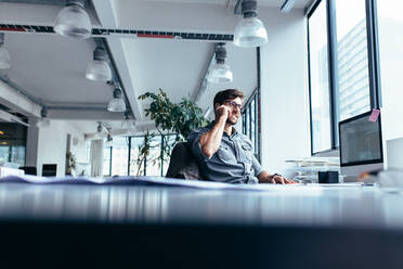 Junger Geschäftsmann, der in einem modernen Büro mit seinem Handy telefoniert. Kaukasischer Mann sitzt an seinem Schreibtisch im Büro. - JLPSF02469