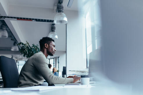 Seitenansicht eines jungen afrikanischen Mannes bei der Arbeit im Büro. Männliche Führungskraft sitzt an seinem Schreibtisch und arbeitet am Computer. - JLPSF02474