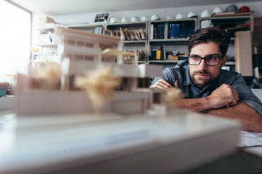 Junger Architekt bei der Arbeit an einem Hausmodell. Männlicher Designer an seinem Schreibtisch, der über neue architektonische Ideen nachdenkt. - JLPSF02490