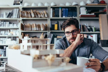 Nachdenklicher männlicher Designer im Büro, der ein Modell mit einer Tasse Kaffee betrachtet. Junger männlicher Architekt, der eine Pause von der Arbeit macht. - JLPSF02491