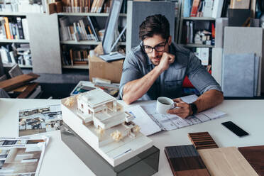 Junger männlicher Architekt in einer Arbeitspause. Kreativer Designer im Büro, der ein Modell mit einer Tasse Kaffee betrachtet. - JLPSF02492