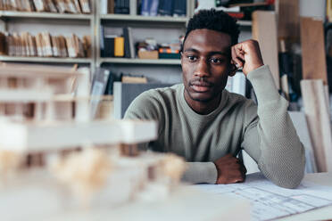 Junger afrikanischer Architekt betrachtet Architekturhausmodell und denkt über neue Ideen nach. Männlicher Designer sitzt im Büro und arbeitet an einem neuen Projekt. - JLPSF02493