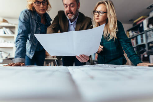 Eine Gruppe von Architekten diskutiert in einem Büro über Baupläne. Ein Geschäftsmann arbeitet mit seinen Kolleginnen und zeigt auf einen Bauplan. - JLPSF02497