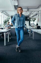 Young female manager standing in office with colleagues working in background. Happy businesswoman looking at camera and smiling. - JLPSF02504