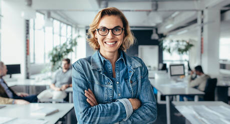 Happy female executive standing with arms crossed. Young businesswoman standing in office. - JLPSF02507