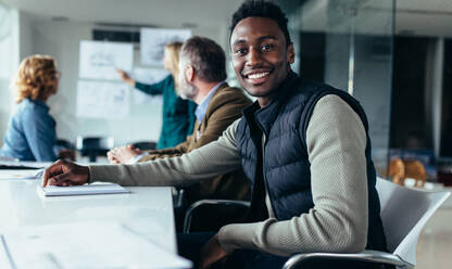 Young businessman sitting in meeting with colleagues presenting in background. Black man sitting in board room. - JLPSF02544