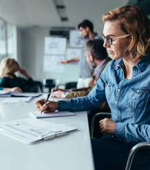 Junge Geschäftsfrau sitzt im Büro mit Geschäftsmann präsentiert im Hintergrund. Weibliche Designer sitzen im Sitzungssaal und schreiben Notizen. - JLPSF02553